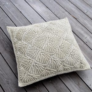 Op de afbeelding: Een beige gehaakt kussen met een decoratief patroon van overlappende cirkels. Het kussen ligt op een houten dek.