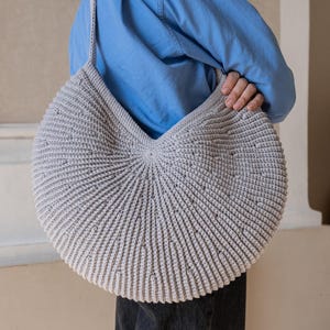 May include: A large, circular, light gray crochet shoulder bag featuring a textured, ribbed radial pattern that radiates from the center. The bag has a single integrated shoulder strap and a consistent, dense stitch construction throughout the design.