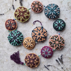 May include: A crocheted garland with a tassel, featuring colourful round ornaments with intricate star-shaped designs. The garland is made with various shades of brown, green, purple, and pink.
