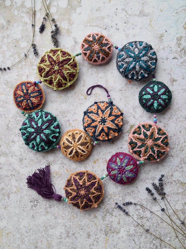 May include: A crocheted garland with a tassel, featuring colourful round ornaments with intricate star-shaped designs. The garland is made with various shades of brown, green, purple, and pink.
