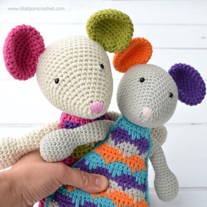 May include: Two crocheted mouse toys, one white with pink, green, and orange ears, and one gray with purple and orange ears. Both mice are wearing colorful striped crocheted dresses.