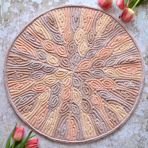 May include: A round crocheted rug with a swirling pattern in shades of peach, beige, and brown. The rug is made with a textured yarn and has a fringed edge.