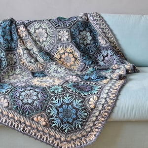 May include: A crochet blanket composed of interconnected hexagonal motifs featuring intricate floral patterns in shades of blue, grey, purple, and soft peach. The edges are finished with a decorative border of repeating petal-like stitches.