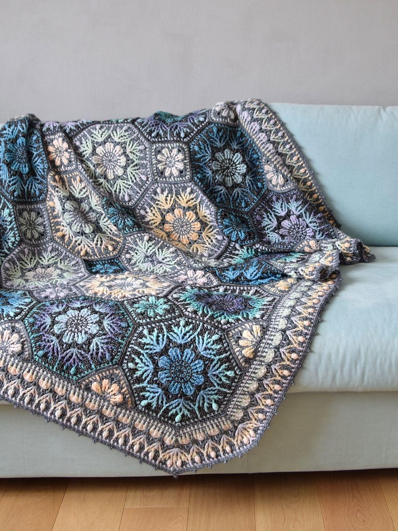May include: A crochet blanket composed of interconnected hexagonal motifs featuring intricate floral patterns in shades of blue, grey, purple, and soft peach. The edges are finished with a decorative border of repeating petal-like stitches.