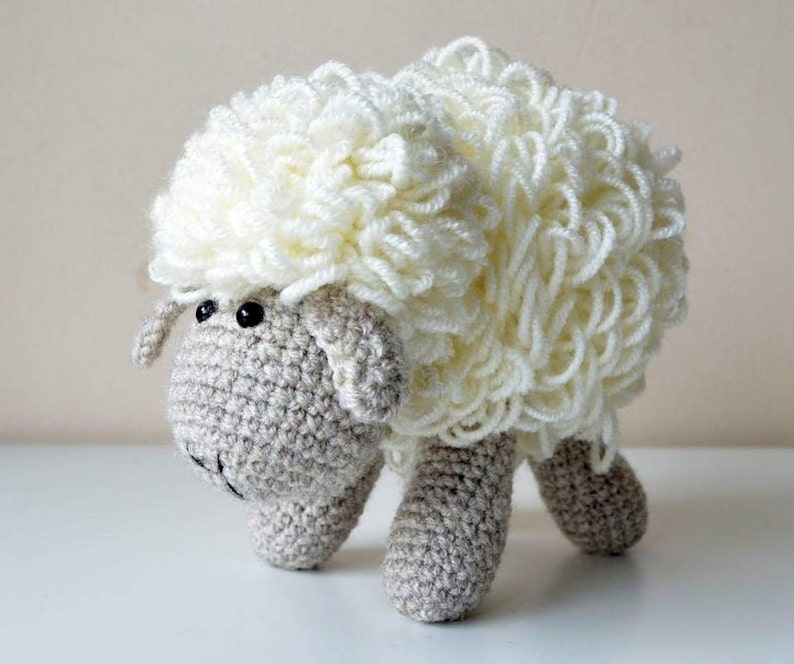 Puede incluir: Un juguete de peluche de oveja blanca de crochet con ojos negros. La oveja tiene un cuerpo de lana blanca esponjosa y una cara y patas grises de crochet.