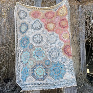 May include: A rectangular crochet blanket featuring a repeating hexagonal motif pattern. The design incorporates a gradient color scheme transitioning from cool blue and grey tones to warm pink, orange, and yellow hues, finished with a decorative border.