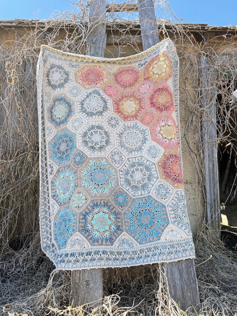 May include: A rectangular crochet blanket featuring a repeating hexagonal motif pattern. The design incorporates a gradient color scheme transitioning from cool blue and grey tones to warm pink, orange, and yellow hues, finished with a decorative border.