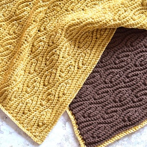 Może przedstawiać: Zbliżenie dwóch szydełkowych koców. Jeden jest w kolorze złotożółtym z teksturowanym, powtarzającym się owalnym wzorem. Drugi koc jest w kolorze ciemnobrązowym z podobnym wzorem. Oba koce mają obramowanie w pasującym kolorze.