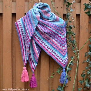 Pode incluir: Um xale triangular de crochê com um padrão geométrico em tons de azul, rosa e roxo. O xale tem borlas nas pontas.