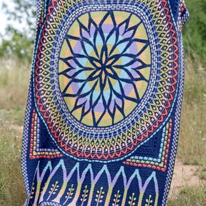 May include: A colourful crocheted blanket with a large circular mandala design in the centre. The mandala features a variety of colours, including blue, green, yellow, orange, and pink. The blanket is bordered by a dark blue crocheted border with a repeating pattern of trees.