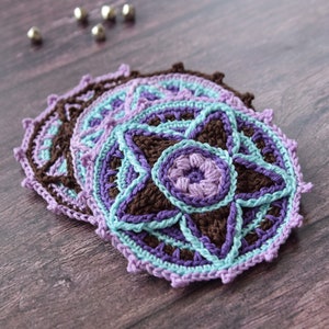 May include: A set of four crocheted coasters in shades of brown, purple, and blue. The coasters are layered on top of each other, with the top coaster featuring a star-shaped design.