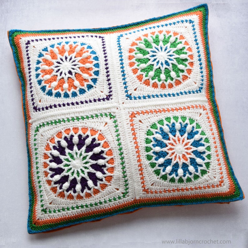 May include: A white crocheted pillow with four colorful circular designs. The designs are in shades of orange, green, blue, and purple. The pillow has a white background with a border of orange, green, and blue.