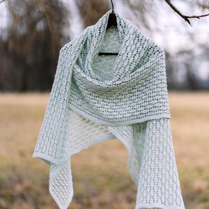 May include: A light green crocheted shawl hanging from a branch. The shawl is made with a delicate open stitch pattern.