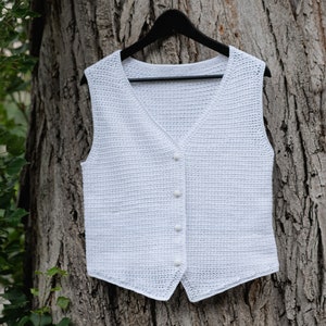 Op de afbeelding: Een witte, gehaakte vest met een V-hals en knoopsluiting, hangend aan een zwarte hanger. Het vest is gemaakt van een textiel met open weefsel. De achtergrond is een boomstam.