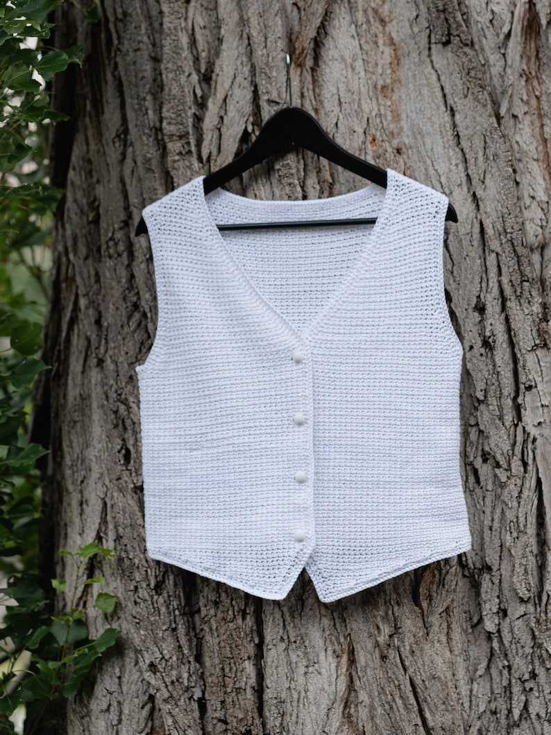 Op de afbeelding: Een witte, gehaakte vest met een V-hals en knoopsluiting, hangend aan een zwarte hanger. Het vest is gemaakt van een textiel met open weefsel. De achtergrond is een boomstam.