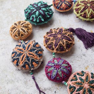 May include: A garland of crocheted round ornaments in various colours and patterns, including teal, brown, purple, and pink. The ornaments are connected by small beads and a purple tassel hangs from one of the ornaments.