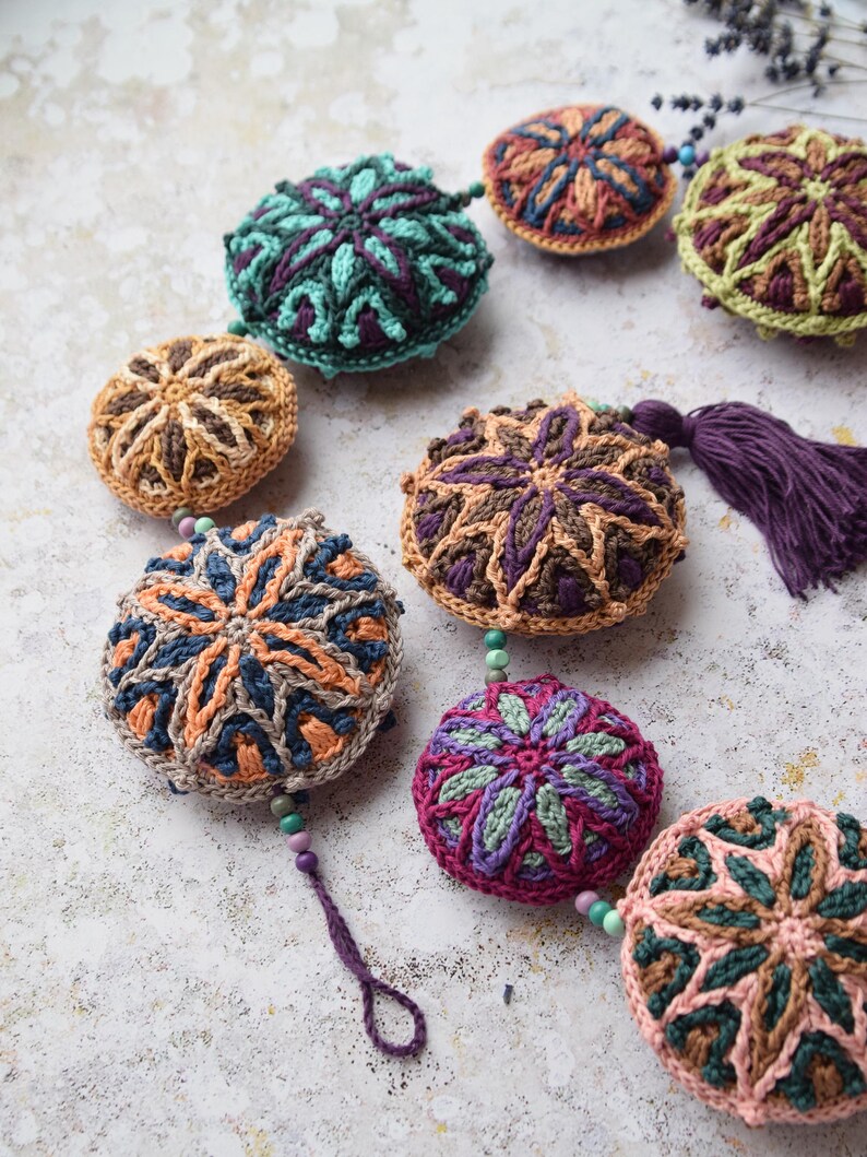 May include: A garland of crocheted round ornaments in various colours and patterns, including teal, brown, purple, and pink. The ornaments are connected by small beads and a purple tassel hangs from one of the ornaments.