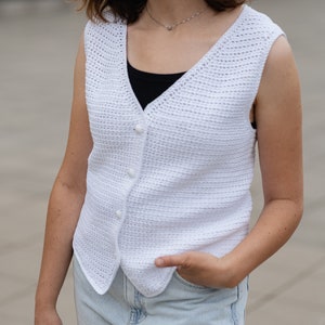 Op de afbeelding: Witte mouwloze vest met knopen. Het vest is gemaakt van een getextureerd, gehaakt materiaal. Het vest heeft een V-hals en wordt over een zwarte top gedragen. De persoon draagt lichtblauwe jeans.