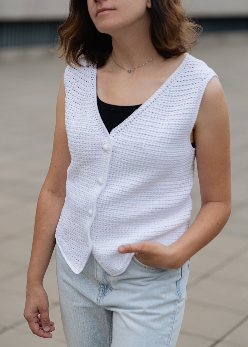 Op de afbeelding: Witte mouwloze vest met knopen. Het vest is gemaakt van een getextureerd, gehaakt materiaal. Het vest heeft een V-hals en wordt over een zwarte top gedragen. De persoon draagt lichtblauwe jeans.