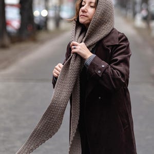 Pode incluir: Uma mulher vestindo um casaco de camur&ccedil;a marrom e um longo cachecol de tric&ocirc; marrom. O cachecol est&aacute; drapeado sobre sua cabe&ccedil;a e amarrado ao redor do pesco&ccedil;o. Ela est&aacute; em p&eacute; em uma rua.