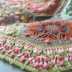 May include: A crochet blanket composed of interconnected hexagonal motifs featuring intricate floral patterns in shades of sage green, soft pink, coral, and cream. The textured yarn work displays detailed stitching, including raised bobbles and petals.