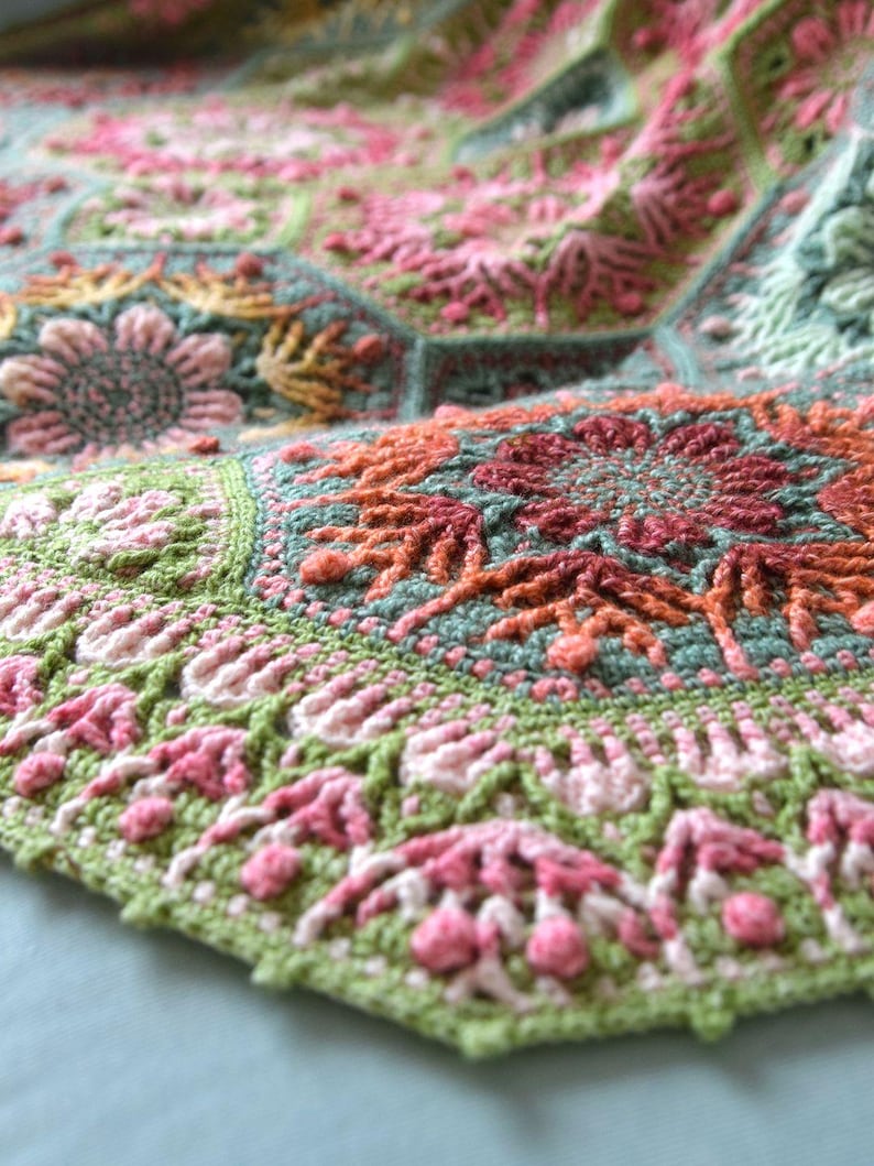 May include: A crochet blanket composed of interconnected hexagonal motifs featuring intricate floral patterns in shades of sage green, soft pink, coral, and cream. The textured yarn work displays detailed stitching, including raised bobbles and petals.