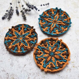 May include: Three crocheted coasters featuring a brown, orange and teal geometric design. The coasters are round and have a raised edge.