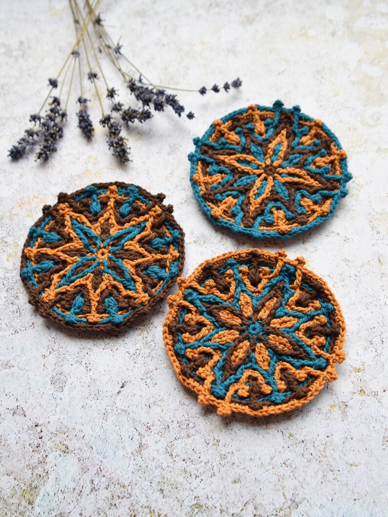 May include: Three crocheted coasters featuring a brown, orange and teal geometric design. The coasters are round and have a raised edge.