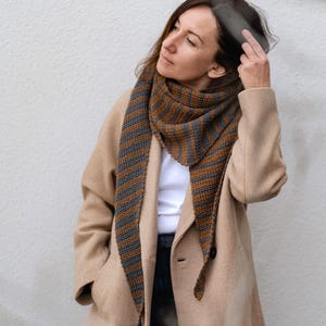 May include: A woman wearing a beige coat with a brown and gray striped scarf. The scarf is tied around her neck and hangs down to her waist.