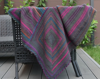 May include: A colorful crocheted blanket with a geometric pattern in shades of gray, pink, orange, and blue. The blanket is draped over a black wicker chair.
