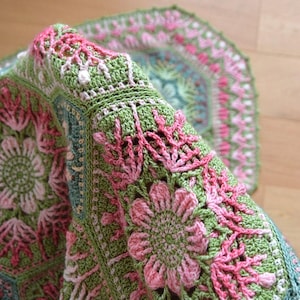 May include: A crochet blanket featuring a repeating hexagonal pattern in shades of sage green, soft pink, and muted teal. The intricate design showcases floral motifs with raised textures and delicate openwork stitches throughout the fabric.