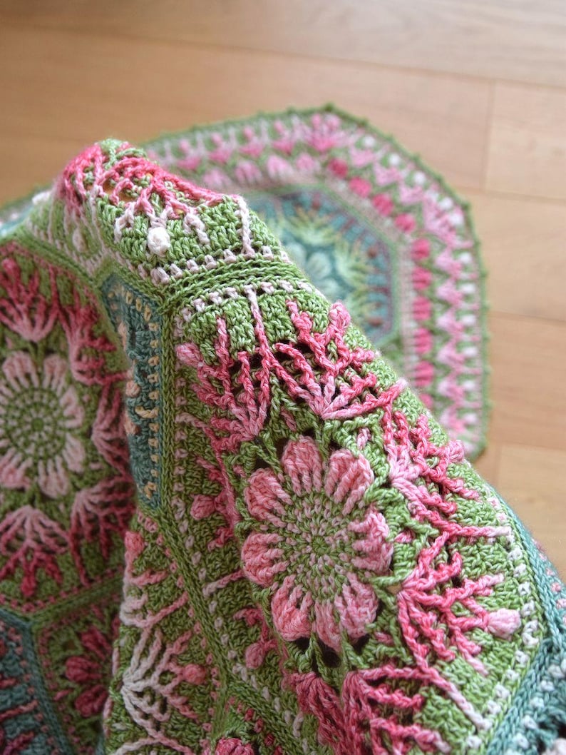 May include: A crochet blanket featuring a repeating hexagonal pattern in shades of sage green, soft pink, and muted teal. The intricate design showcases floral motifs with raised textures and delicate openwork stitches throughout the fabric.