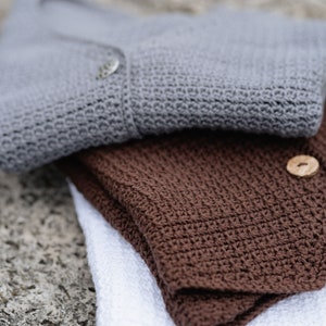 Op de afbeelding: Close-up van drie gevouwen gehaakte items. Een grijze cardigan met een knoop ligt bovenop, gevolgd door een gevouwen bruin stuk en een wit stuk onderaan. De items zijn gemaakt van een getextureerd, gebreid materiaal.