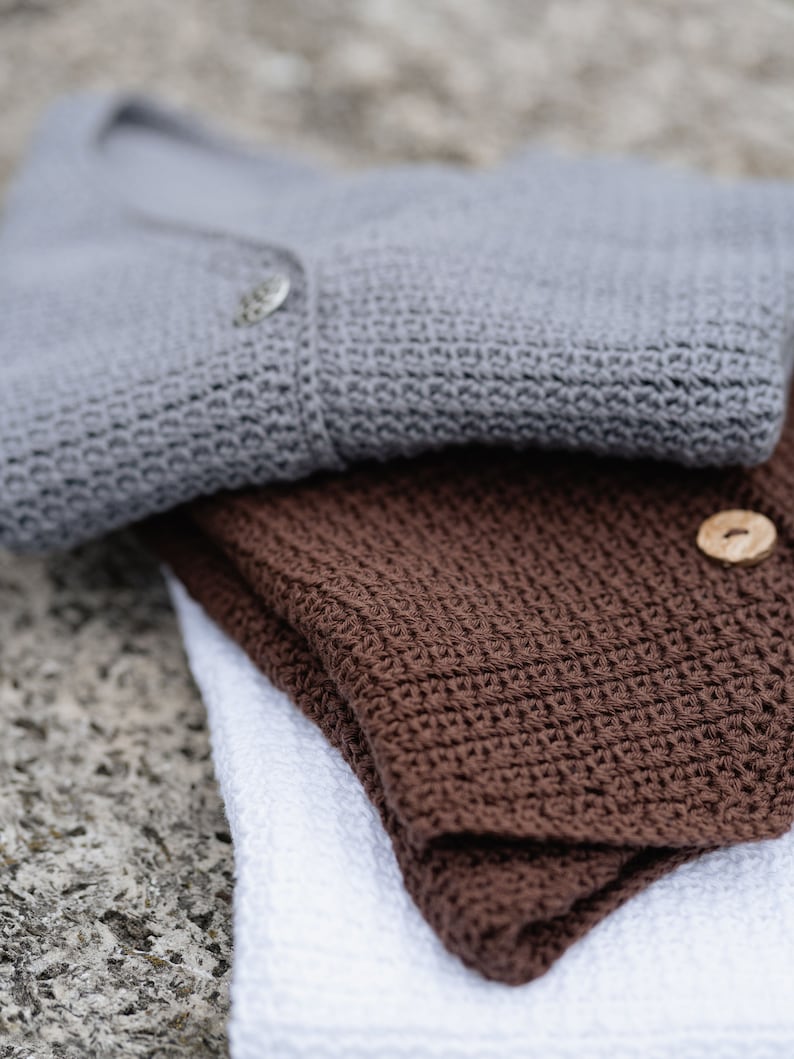 Op de afbeelding: Close-up van drie gevouwen gehaakte items. Een grijze cardigan met een knoop ligt bovenop, gevolgd door een gevouwen bruin stuk en een wit stuk onderaan. De items zijn gemaakt van een getextureerd, gebreid materiaal.