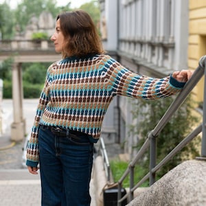以下が含まれることがあります： 茶色、ターコイズ、白の模様のセーター。セーターはクルーネックで、ターコイズのカフスが付いた長袖です。濃い青色のジーンズと黒いベルトを着用しています。背景には建物と階段が写っています。