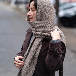 Pode incluir: Uma echarpe com capuz de croch&ecirc; marrom com um padr&atilde;o texturizado. A echarpe est&aacute; sendo usada por uma pessoa em uma jaqueta marrom.