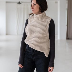 Op de afbeelding: Een beige, geribbelde gebreide coltrui vest met drie knopen aan de zijkant. Het vest wordt gedragen over een donkergrijs shirt met lange mouwen en gecombineerd met een zwarte jeans. De outfit is eenvoudig en stijlvol.