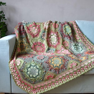 May include: A handmade crochet blanket featuring a complex arrangement of hexagonal motifs. The design incorporates floral patterns in shades of sage green, dusty pink, soft yellow, and cream, finished with a decorative scalloped border edge.