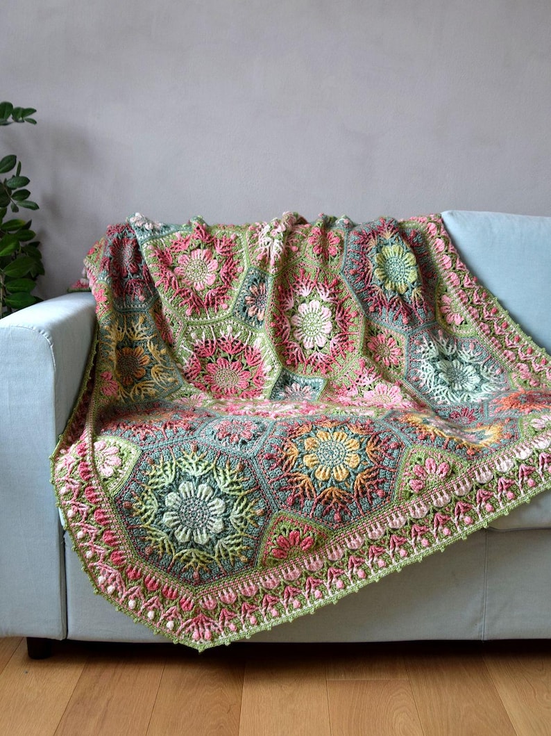 May include: A handmade crochet blanket featuring a complex arrangement of hexagonal motifs. The design incorporates floral patterns in shades of sage green, dusty pink, soft yellow, and cream, finished with a decorative scalloped border edge.