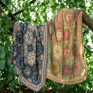 May include: A large, rectangular crochet blanket featuring a complex pattern of interconnected hexagonal motifs. The left side displays dark charcoal, blue, and cream tones, while the right side transitions into warm shades of olive, pink, and gold.