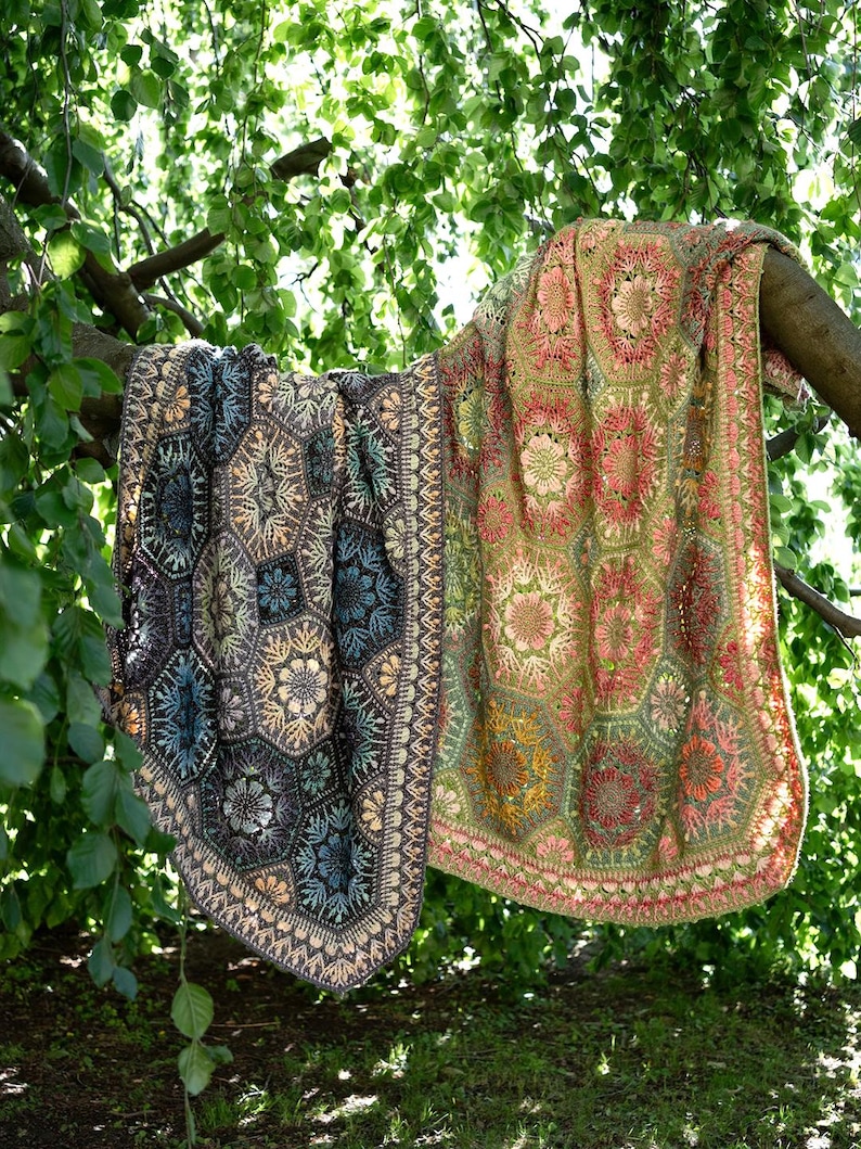 May include: A large, rectangular crochet blanket featuring a complex pattern of interconnected hexagonal motifs. The left side displays dark charcoal, blue, and cream tones, while the right side transitions into warm shades of olive, pink, and gold.