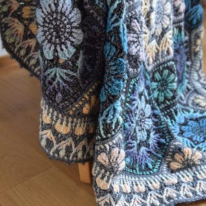 May include: A crochet blanket featuring a complex arrangement of hexagonal motifs in shades of charcoal gray, light blue, teal, and soft peach. The design incorporates intricate floral patterns with raised textures and detailed stitching throughout.