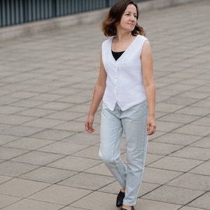 Op de afbeelding: Een witte vest met knopen en een textuurpatroon. De vest wordt gedragen over een zwarte top en gecombineerd met lichtblauwe jeans en zwarte schoenen. De persoon loopt op een grijze tegelvloer.
