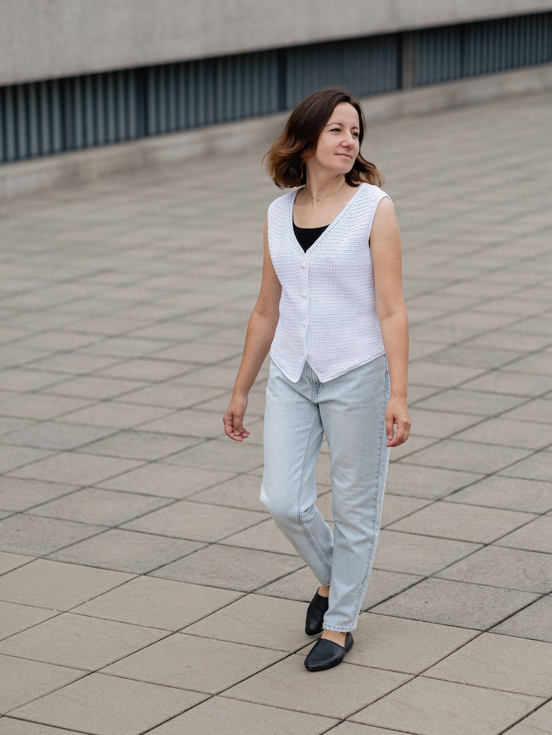 Op de afbeelding: Een witte vest met knopen en een textuurpatroon. De vest wordt gedragen over een zwarte top en gecombineerd met lichtblauwe jeans en zwarte schoenen. De persoon loopt op een grijze tegelvloer.