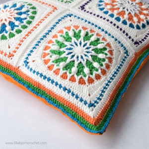May include: A white crocheted pillow with a colorful geometric pattern. The pillow has a border of orange, green, and blue yarn.
