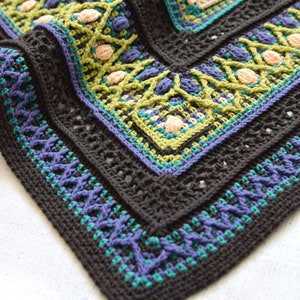 Puede incluir: Primer plano de una manta de ganchillo con un esquema de color negro, verde, morado y crema. La manta tiene un patrón geométrico y un borde texturizado.