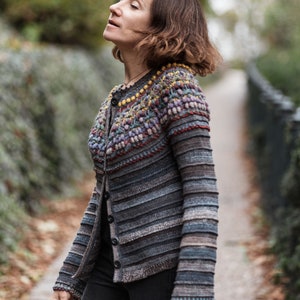 Puede incluir: Una mujer lleva una chaqueta de punto de rayas, de ganchillo, con un yugo de colores y estampado. La chaqueta es de color gris oscuro y marrón con un diseño de ganchillo texturizado. La mujer está de pie en un parque con árboles y vegetación en el fondo.