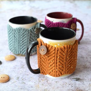 May include: Three ceramic mugs with knitted cozy covers. The mugs are in different colors: blue, orange, and purple. The cozies are knitted in a chevron pattern.