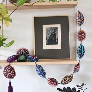 May include: A colourful crocheted garland with purple, pink, blue and green accents hangs on a wooden shelf. The garland is made of crocheted balls with intricate patterns. A framed black and white print hangs on the shelf above the garland.