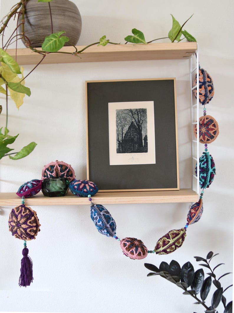 May include: A colourful crocheted garland with purple, pink, blue and green accents hangs on a wooden shelf. The garland is made of crocheted balls with intricate patterns. A framed black and white print hangs on the shelf above the garland.